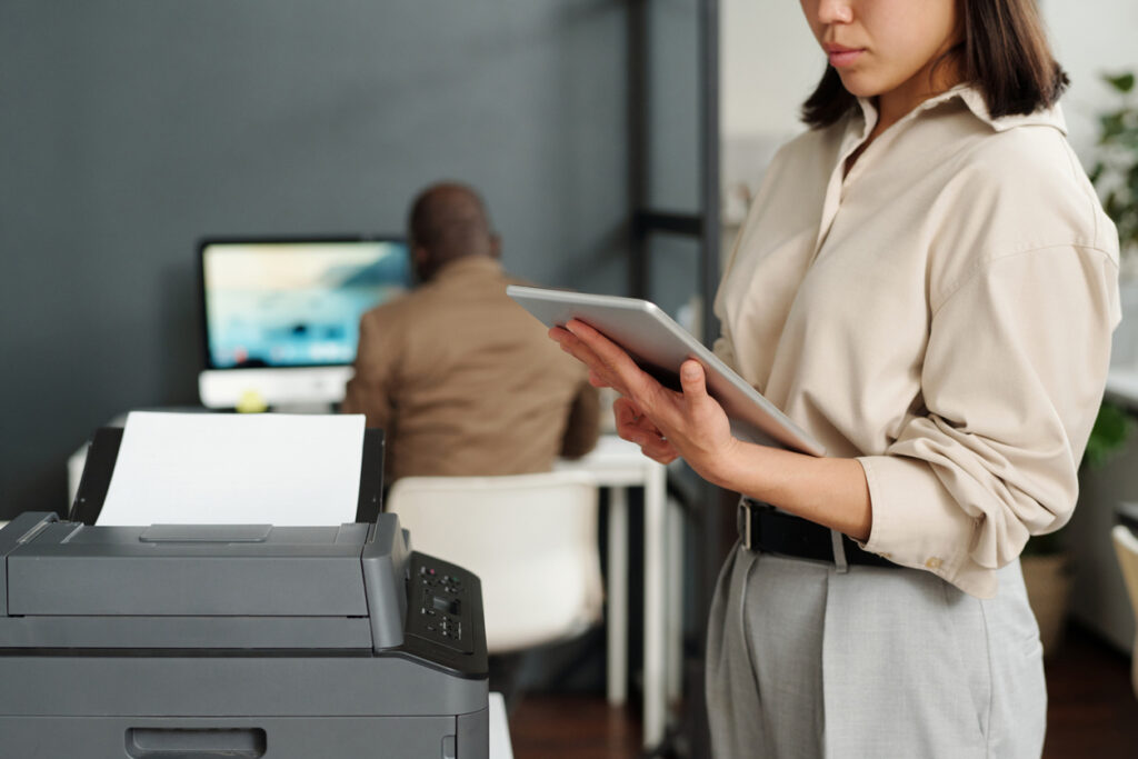 Focus on tablet held by businesswoman computing in office while standing by a printer